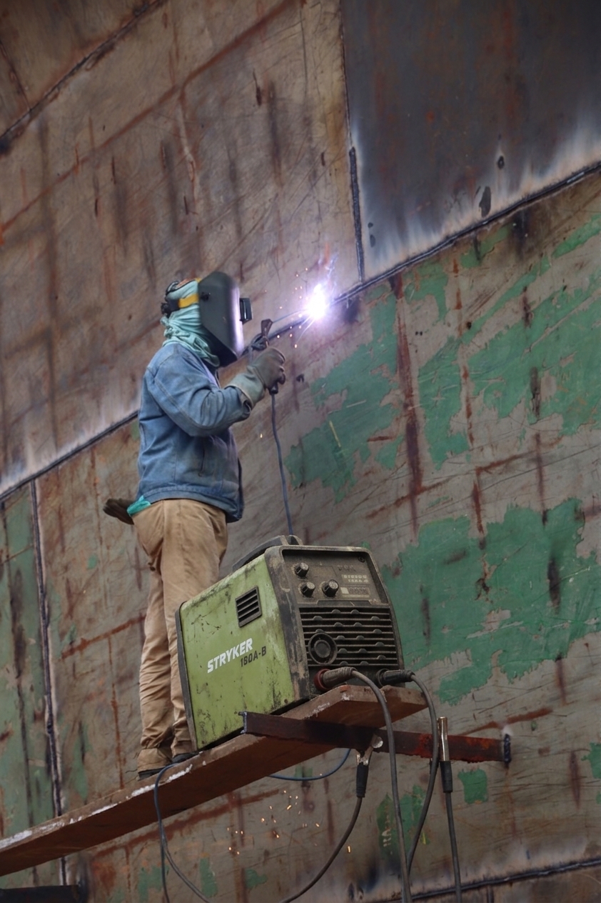 Welder on a raised plank using a Stryker 150A-B to weld a large steel hull